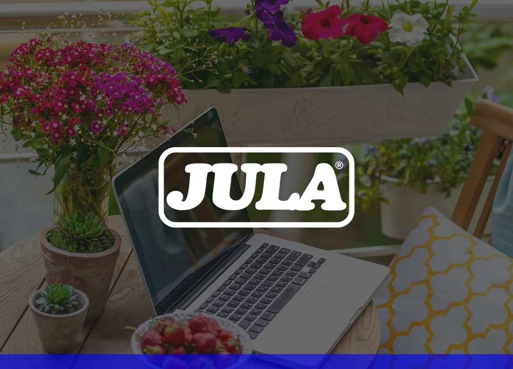 Laptop auf einem Tisch im Freien, umgeben von Pflanzen und einem Kissen, mit dem Logo von JULA.