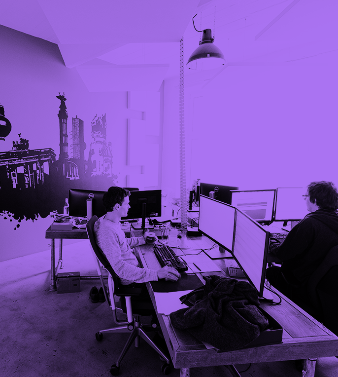 a couple of men sitting at a desk with computers and a purple wall